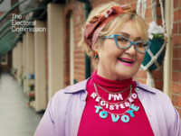 A woman is standing outside her flat, looking happy. She is wearing a necklace with “I’m Registered To Vote” spelt out on it.