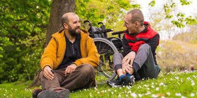 Image of 2 men sitting on green grass with an empty wheelchair in the background