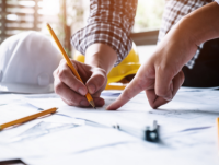 Image of hands drawing and pointing on engineering plans with two hard hats in the background